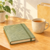 木漏れ日の差す机に置かれた、緑色の閉じたノートとお茶のカップの水彩イラスト